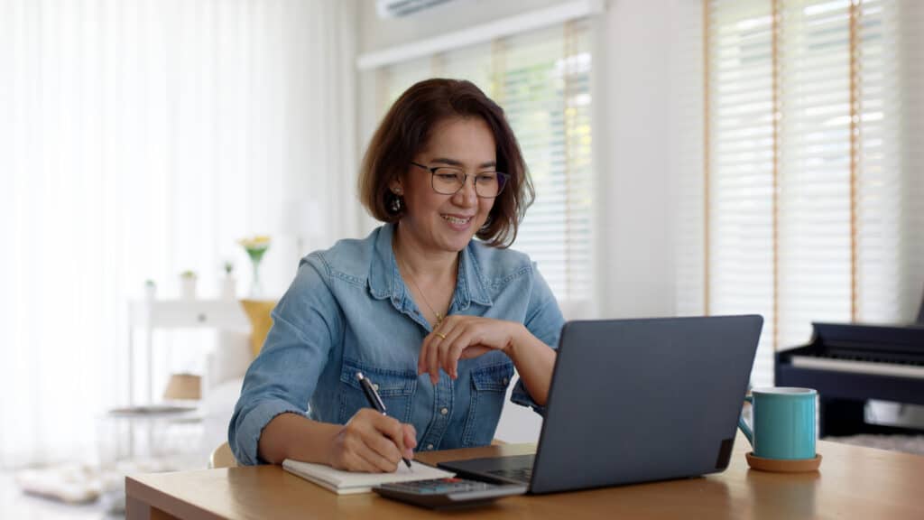 Frau arbeitet am Schreibtisch mit Laptop, Taschenrechner, Notizblock und einer Tasse Kaffee