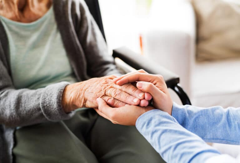 Eine junge Frau hält die Hände einer pflegebedürftigen Frau während eines Hausbesuchs.